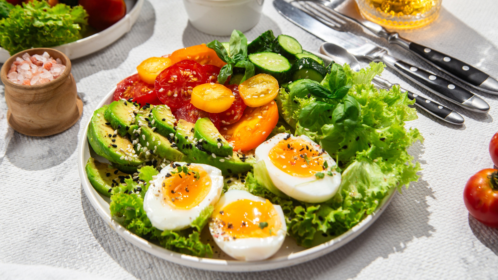 A breakfast plate with sliced avocado, soft-boiled eggs, cherry tomatoes, lettuce, and cucumber in natural light